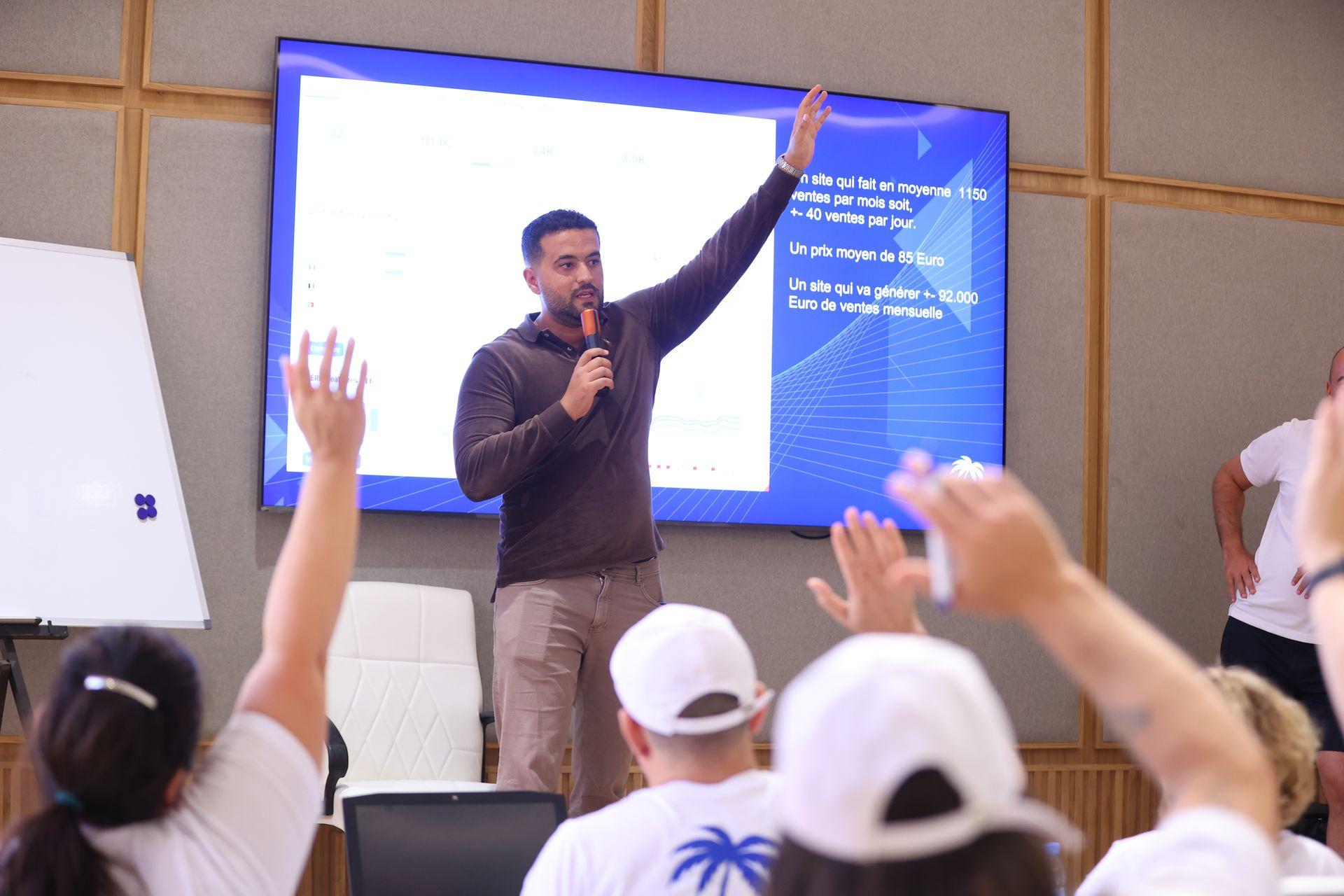 Man giving a presentation with microphone; audience raising hands; large screen displays text and chart.